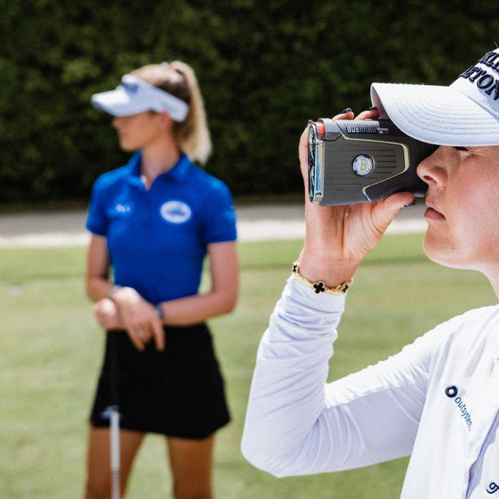 A golfer wearing a visor with a golf rangefinder to her face with another golfer in the background on the golf course