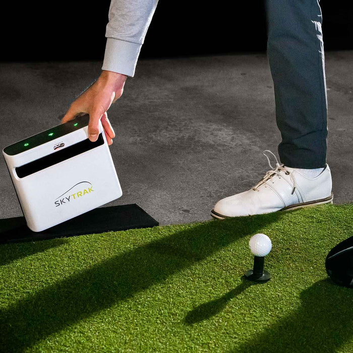 man placing skytrak+ golf simulator and golf launch monitor on the ground next to a golf mat with a golf ball and golf tee sitting on it