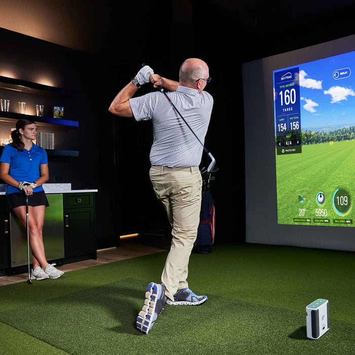 man hitting a golf ball with a golf club in an indoor home skytrak+ golf simulator studio golf with a package of golf mat, impact screen, projector while a young woman golfer looks on