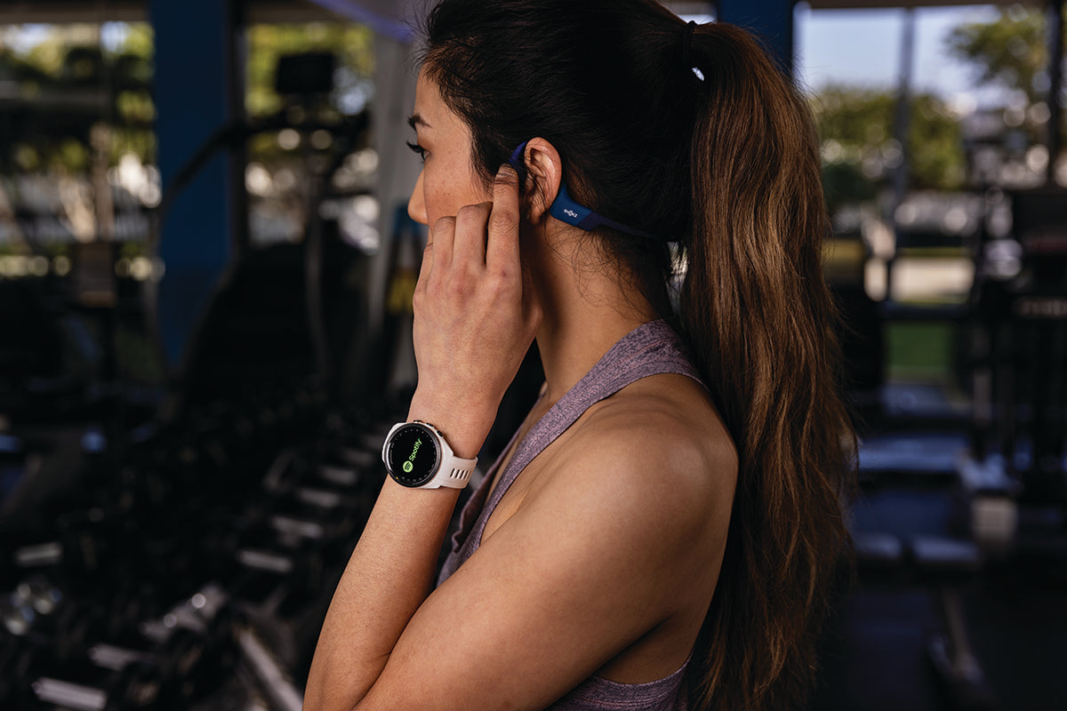 A woman in the gym adjusting her wireless ear buds while wearing a 42 mm Garmin Approach S70 golf watch with the Spotify app on the display