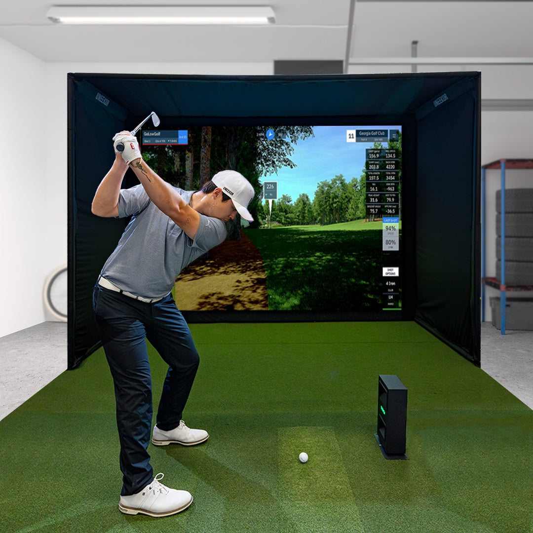 A man with golf club raised in his backswing in a garage golf simulator with a Uneekor Eye Mini Lite launch monitor, golf hitting mat, enclosure and impact screen with golf sim software on the screen