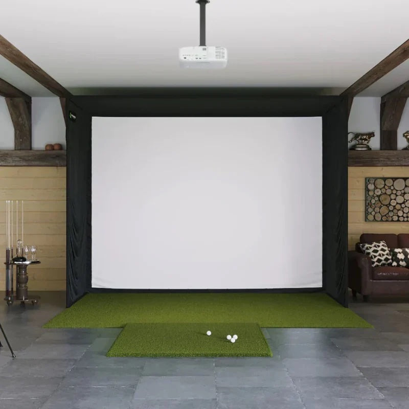 Indoor golf simulator setup with projector, green mat and white screen in a room.