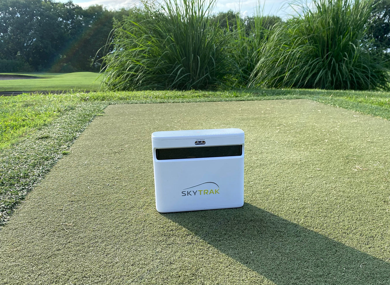 The SkyTrak+ unit on a golf range in front of tall grass and trees