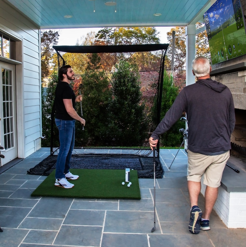 Two guys looking at SkyTrak+ software on a flatscreen TV in an outdoor golf simulator setup with golf hitting net, golf practice mat and SkyTrak+ golf launch monitor