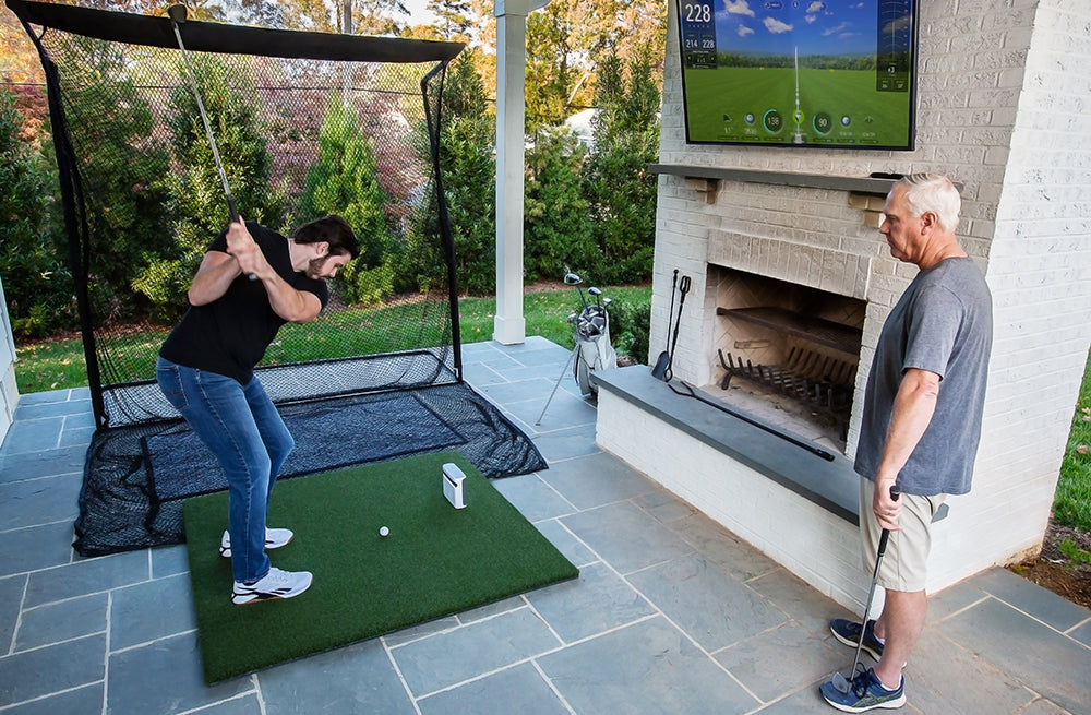 A golfer hitting into a golf practice net using a SkyTrak+ golf launch monitor on a hitting mat with simulation on a mounted flatscreen TV while another man watches