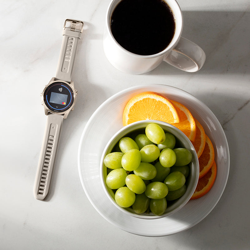 A Garmin fenix 7S laid flat on a table with Garmin Pay on the display next to a cup of coffee and a bowl of grapes on a plate of oranges